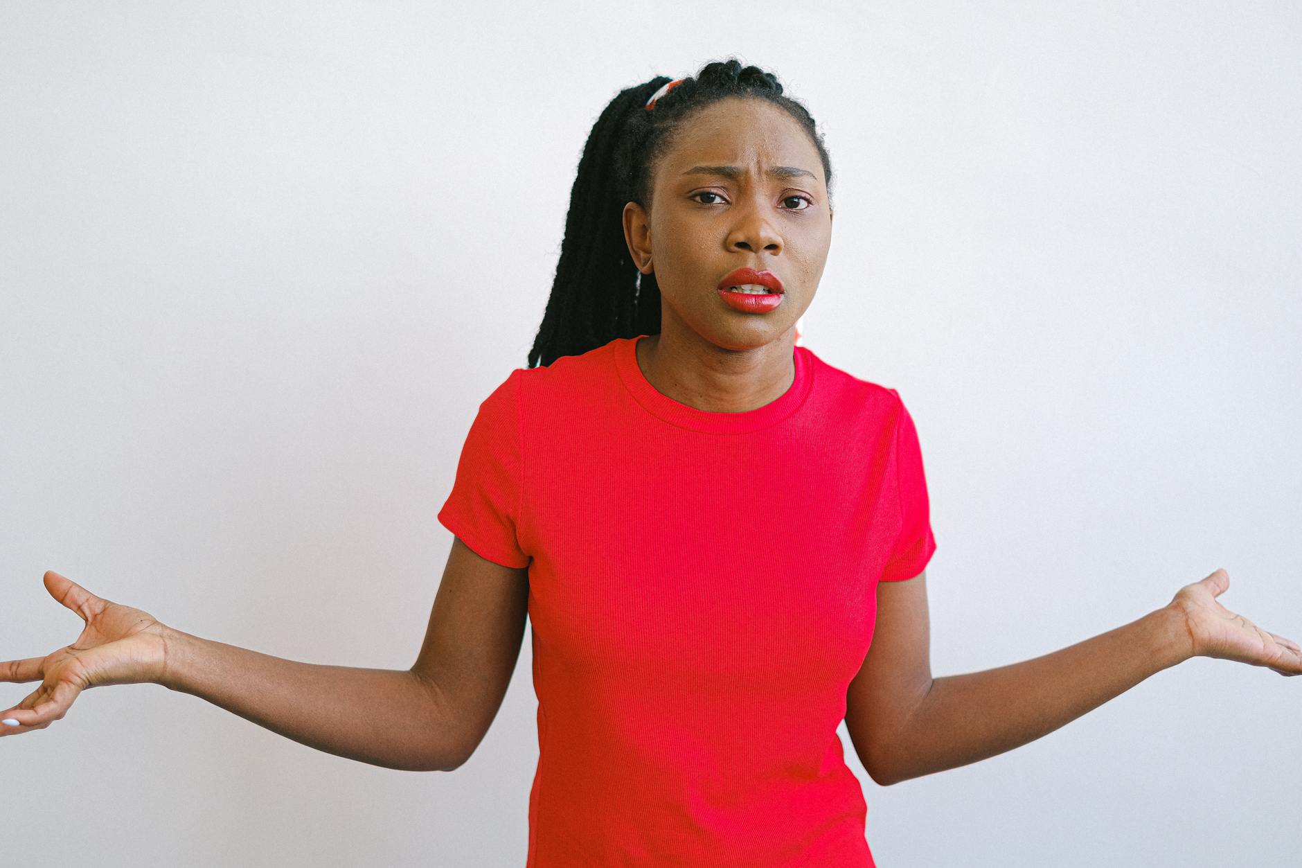 Image of a woman with a red shirt and lipstick looking confused at the camera with her arms at her side in shrugging motion.